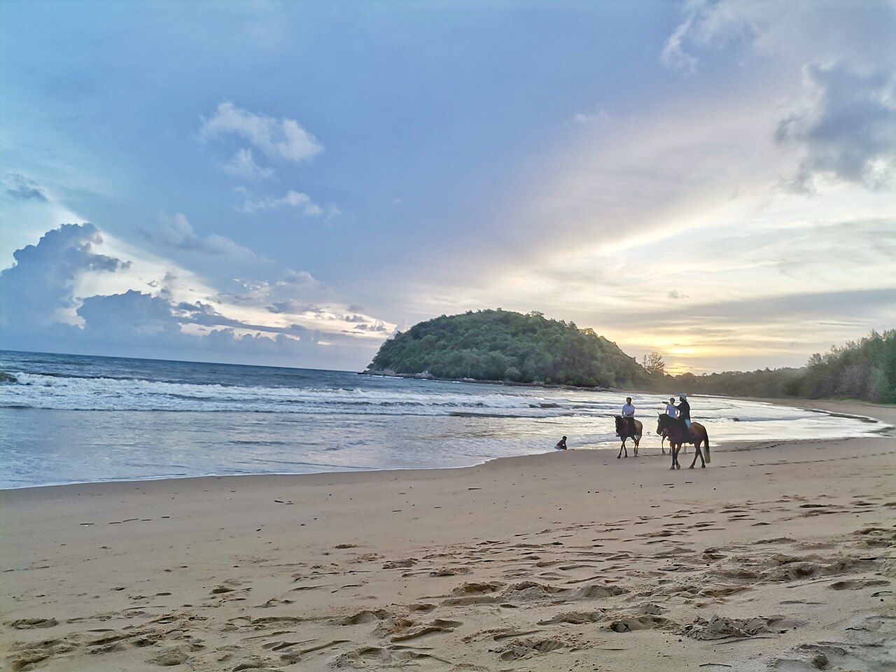 หาดลายัน ภูเก็ต ยามเย็น มีคนขี่ม้าเลียบชายหาด และเนินเขาเขียวริมทะเลด้านหลัง